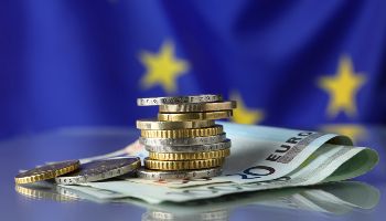 Ein Euro-Münzenstapel liegt auf Euro-Geldscheinen vor einer EU-Flagge