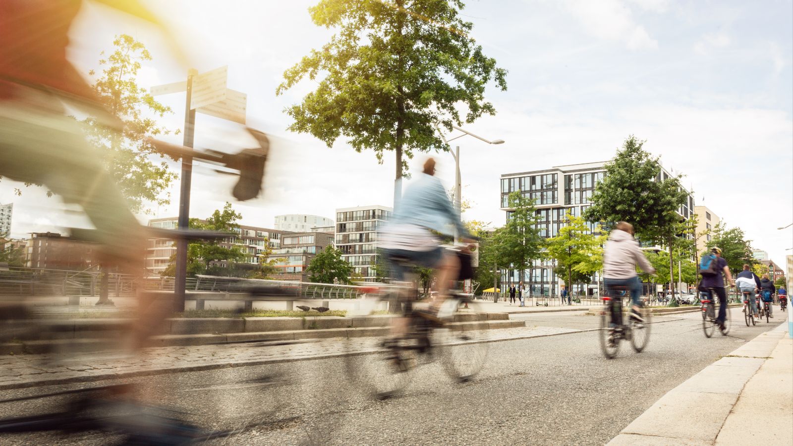 Mehrere Fahrradfahrer fahren hintereinander auf einem Fahrradweg in einer Stadt. Im Hintergrund sind Häuser und Bäume zu sehen.