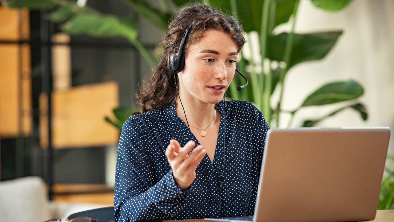 Eine Frau in gepunkteter Bluse sitzt mit einem Headset vor einem Laptop. Sie spricht und gestikuliert. Im unscharfen Hintergrund steht eine große Pflanze.