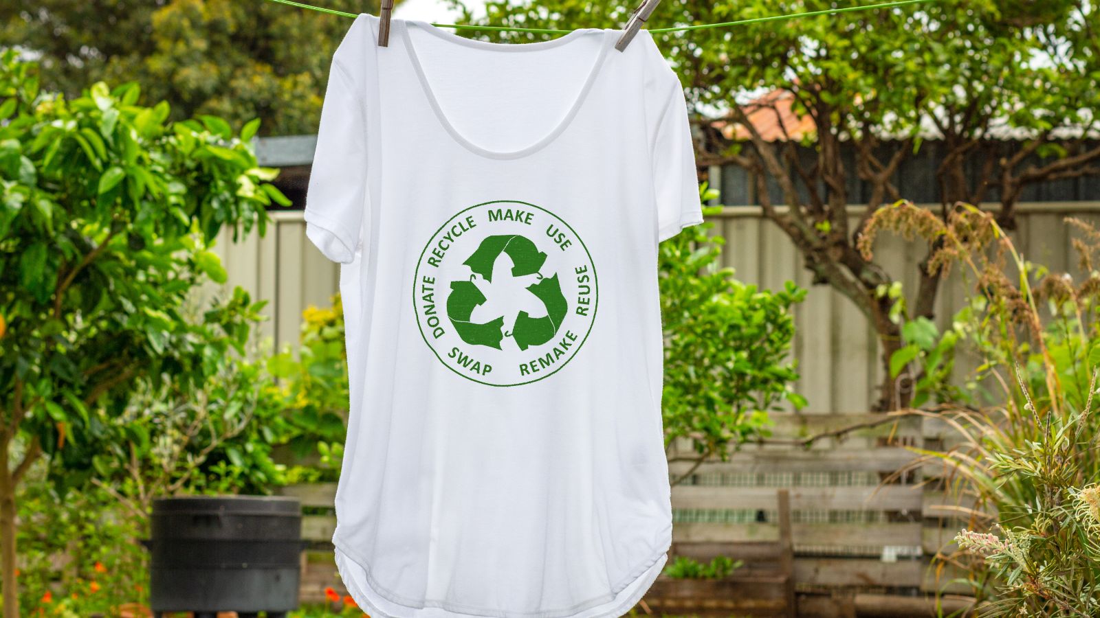 Ein weißes T-Shirt mit einer grünen Abbildung eines Recyclingkreislaufs hängt auf einer Leine in einem grünen Garten.