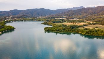 Ein glatter See erstreckt sich zu Füßen einer mit Wäldern bedeckten Gebirgslandschaft. Das Seeufer bildet ein grünes Band aus Büschen und Bäumen.
