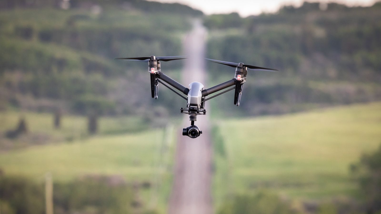 Drone fliegt über eine Landschaft. Im unscharfen Hintergrund ist eine Landstraße erkennbar, welche bergauf führt und von Wald und Wiesen umgeben ist.