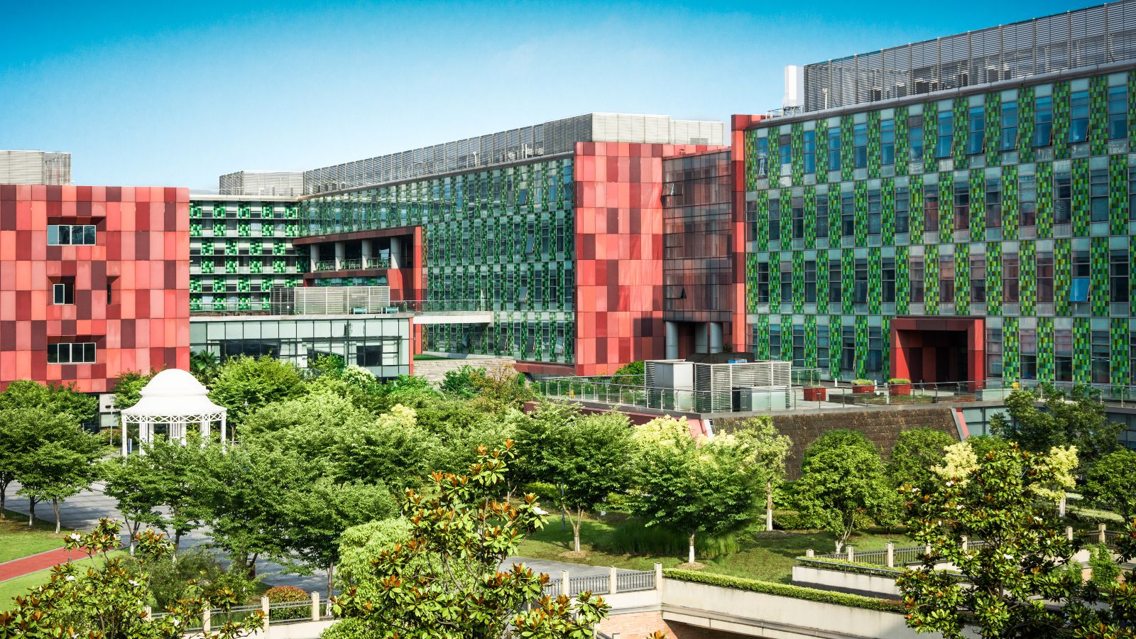 Von Bürogebäuden eingerahmte Grünfläche mit Bäumen, Wegen und einem Pavillon. Im Hintergrund ist blauer Himmel zu sehen.