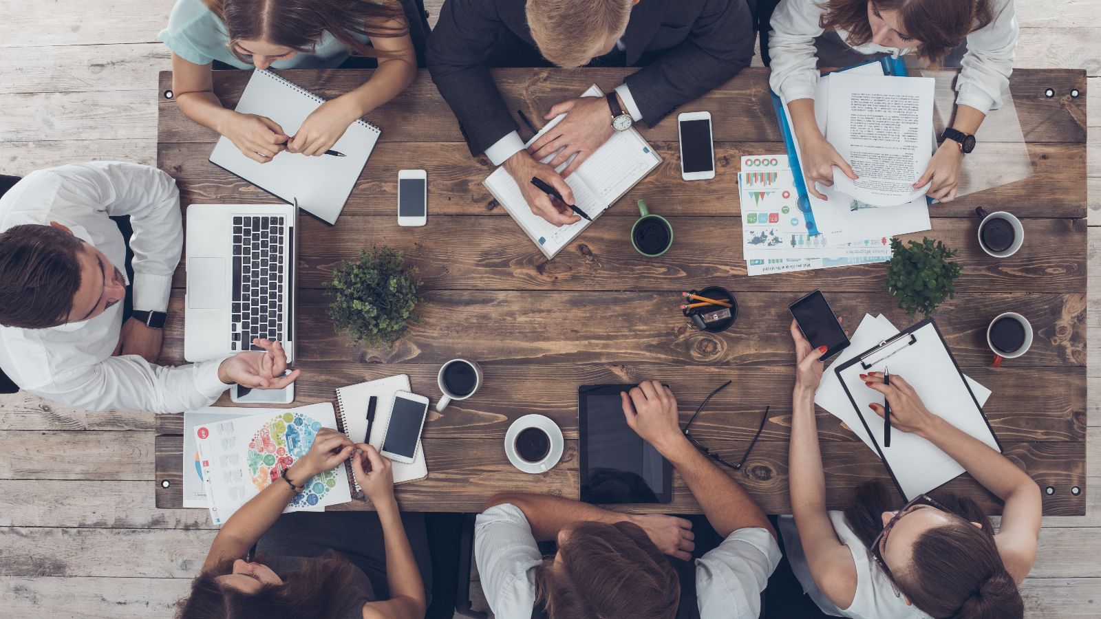 Draufsicht auf einen Tisch mit Unterlagen, Laptops, Tablets, Smartphones und Kaffeetassen. Um den Tisch sitzen sieben Personen und diskutieren.