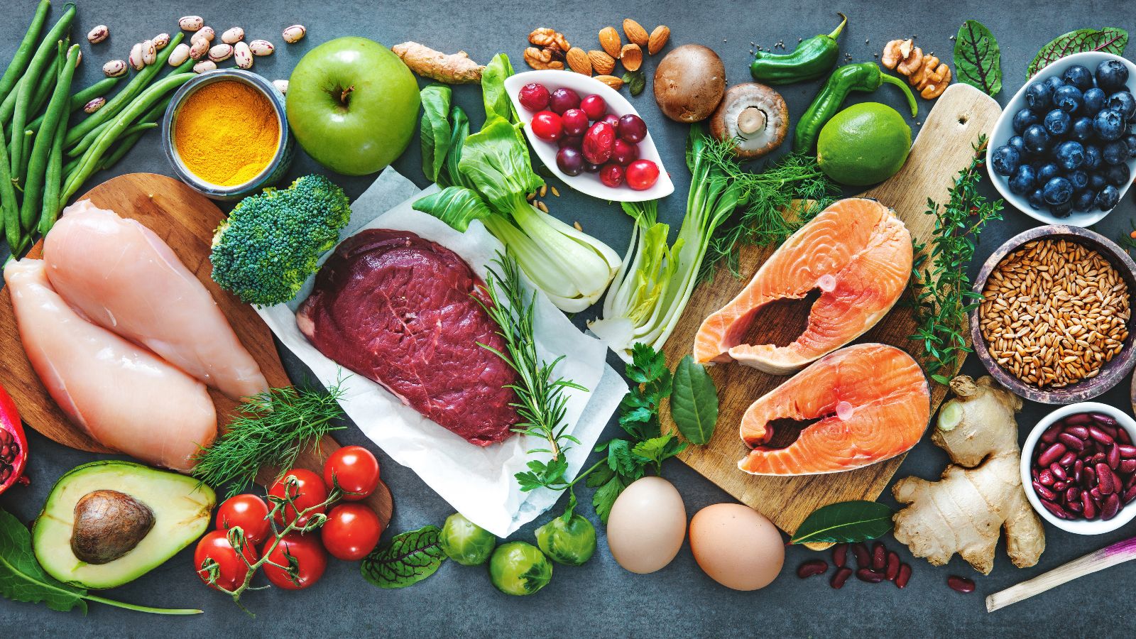 Eine dunkle Arbeitsfläche ist flächendeckend mit diversen frischen Lebensmitteln bedeckt.  Die Lebensmittelauswahl erstreckt sich von Obst und Gemüse neben Fleisch, Fisch und Eiern.