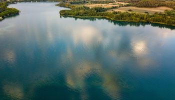 Ein glatter See erstreckt sich zu Füßen einer mit Wäldern bedeckten Gebirgslandschaft. Das Seeufer bildet ein grünes Band aus Büschen und Bäumen. Im Umland erstrecken sich eine Siedlung und vereinzelte Felder.